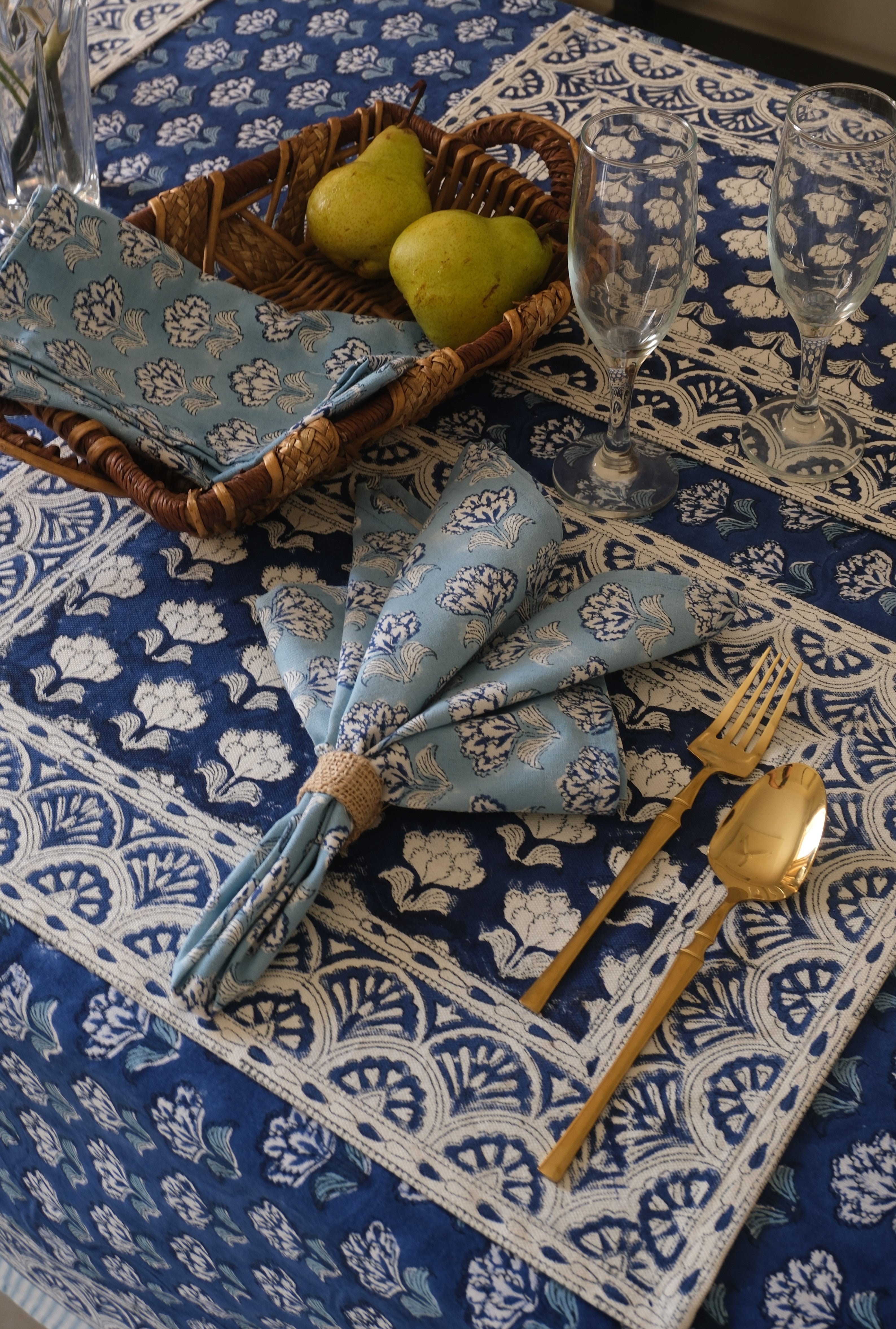 Table setting with blue patterned napkins, gold cutlery, and pears on a blue floral tablecloth.