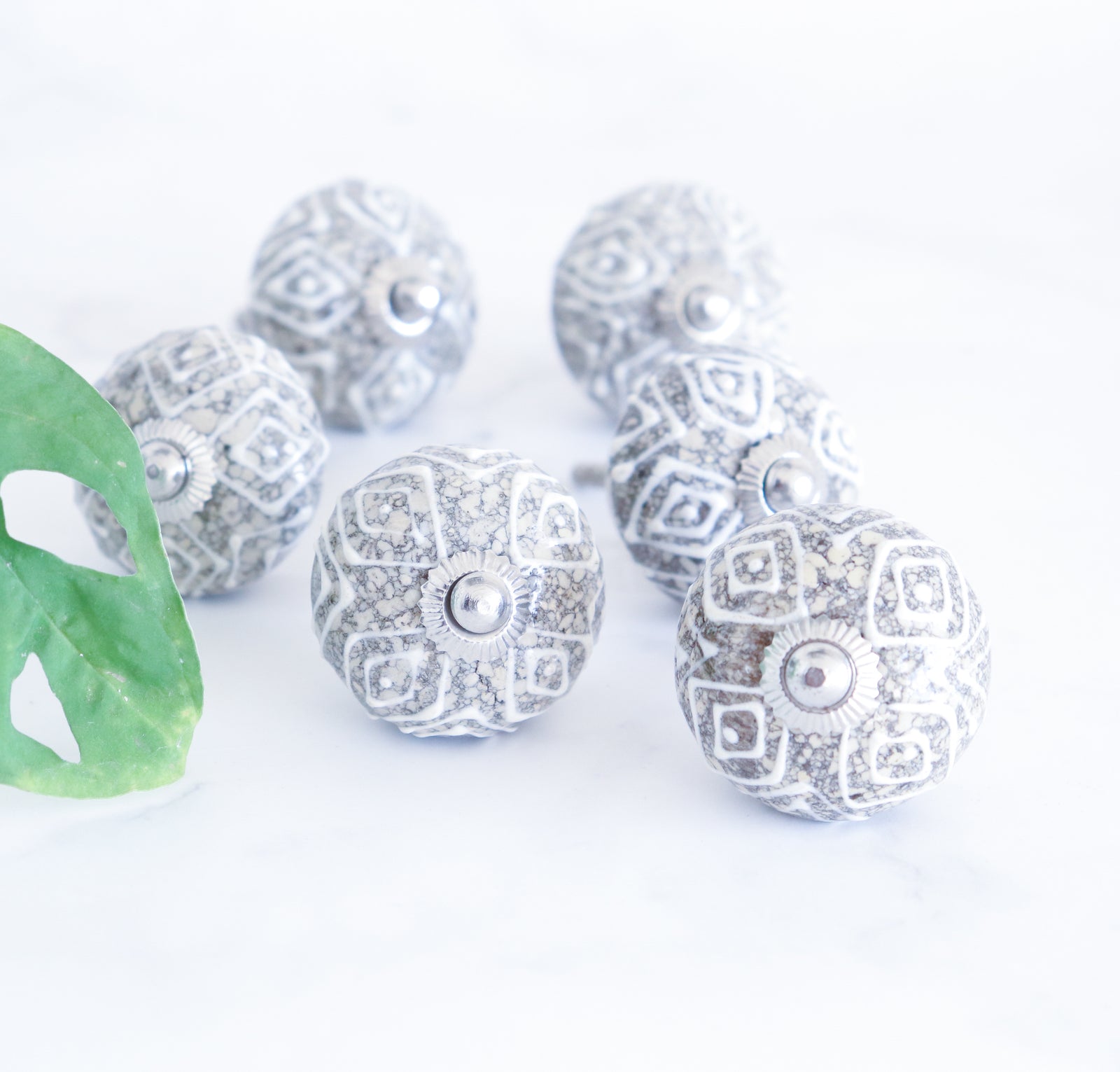 A set of 6 ceramic knobs with a geometric pattern and metal accents, displayed on a white surface with a green leaf nearby.